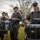 A group of military personnel in Marine Corps MARPAT camouflage uniforms are playing instruments as part of a marching band outdoors. At least three drummers are visible playing Pearl brand snare drums, with brass instrument players visible in the background. The setting appears to be a grassy area with trees and an overcast sky.