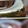 A close-up photograph of a brass trombone bell, with a distorted reflection visible on its polished surface showing a musician wearing a camouflage cap playing an instrument. The reflection captures a tree, sky, and surrounding environment in the curved mirror-like surface of the instrument. The image demonstrates creative photography using the instrument's reflective bell as a compositional element.