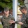 Two U.S. Marines in MARPAT woodland camouflage uniforms stand near a flagpole with a brass bell visible at the top. A senior Marine (identifiable by a rank insignia pin on his collar) appears to be addressing or instructing a younger Marine whose name tape reads 'KAUTZER'. This appears to be a formal military ceremony or instructional moment on a base parade ground.