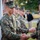 A group of military personnel in MARPAT (Marine pattern) camouflage uniforms are participating in what appears to be a formal ceremony or recognition event outdoors. The foreground shows a handshake between two service members, suggesting an award presentation, promotion, or official greeting. Several other uniformed Marines stand in line in the background, consistent with a formation or receiving line.