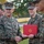 A formal U.S. Marine Corps ceremony is taking place outdoors, where a senior officer identified by name tape as 'DONOVAN' is shaking hands with another Marine while presenting a red certificate folder bearing the Marine Corps emblem. Multiple Marines in MARPAT digital camouflage uniforms are present, suggesting an awards or recognition ceremony. The atmosphere appears positive, with visible smiles among the participants.