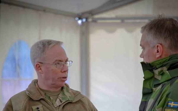 Two military personnel are engaged in conversation inside a tent or temporary structure. One is a U.S. Army officer wearing an olive/tan fleece with the name tape 'BRADY' and two-star rank insignia, and the other is wearing Swedish Armed Forces camouflage with a 'SWEDEN' patch and Swedish national emblem. The interaction appears to be a bilateral military meeting or exercise.