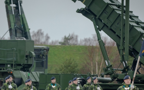 A military ceremonial formation is taking place in front of what appears to be Patriot missile defense system launchers painted in dark green. Soldiers in camouflage uniforms with white gloves are standing at attention, with a standard-bearer in the foreground carrying a blue flag bearing a yellow/gold three-crown emblem consistent with Swedish military insignia. The setting appears to be an outdoor military installation or parade ground under overcast skies.