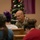 A military service member in camouflage uniform is engaged in conversation with civilians at what appears to be an indoor gathering during the Christmas holiday season. A decorated Christmas tree is visible in the background, suggesting a holiday event or community outreach function. The soldier appears to be smiling and interacting warmly with attendees seated around tables.