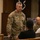 A U.S. Army soldier with the name tape 'HAYNIE' and rank insignia visible on his OCP (Operational Camouflage Pattern) uniform is engaged in conversation with seated civilians in what appears to be an indoor venue or auditorium. A second soldier in the background also wears OCP uniform. The setting suggests a community engagement event or briefing.