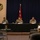 A U.S. Army Hawaii panel discussion or town hall meeting is taking place, with five individuals seated behind a black-draped table bearing the U.S. Army Hawaii logo and 'Forward Together' motto. A central figure in military uniform is speaking into a microphone, flanked by four others including one civilian in a Hawaiian shirt. A one-star general officer flag (Brigadier General) is displayed behind the panel, and Christmas wreaths are visible on the walls.