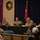 A U.S. Army panel discussion or town hall meeting is taking place at what appears to be a military installation. Five individuals are seated at a long table with a U.S. Army banner, including four uniformed military personnel and one civilian, with an audience seated in rows facing them. A red general officer flag with two stars is visible in the background, along with holiday decorations including a wreath.