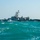 A gray military patrol vessel is operating in choppy turquoise-green waters alongside what appears to be a partially submerged or capsized semi-submersible vessel with a dark camouflage pattern. The scene suggests a maritime interdiction or law enforcement operation at sea. Personnel are visible on the deck of the military vessel.