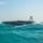 A high-performance speedboat with a dark geometric camouflage pattern is cutting through choppy turquoise waters. The vessel appears to be an unmanned or remotely operated surface vessel (USV) given the absence of visible crew and the presence of sensor/antenna mast equipment. The image is taken in clear daylight conditions with a light blue sky in the background.