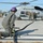 FARP Fleet Battle Problem 21-3 Military personnel are conducting a hot refueling operation on a U.S. Navy MH-60R Seahawk helicopter assigned to HSM-72 (Helicopter Maritime Strike Squadron 72), tail number 704, with the nickname 'Proud Warriors' visible on the fuselage. A crew member in the foreground kneels on the tarmac managing a fuel hose connected to a fuel control valve, while three other personnel stand near the helicopter. The rotors appear to be spinning, indicating a hot refueling (HIFR or FARP) procedure.