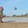 FARP Fleet Battle Problem 21-3 A U.S. Marine in camouflage utilities holds a large red flag on a concrete airstrip or landing zone while a military helicopter, appearing to be a UH-60 Black Hawk or similar utility helicopter, approaches for landing. The scene takes place at what appears to be a military airfield or forward operating area, with trees, ground support equipment, and other personnel visible in the background. The red flag is likely being used as a wind indicator or landing signal for the incoming aircraft.