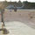 FARP Fleet Battle Problem 21-3 Military personnel in olive drab flight suits are conducting forward area refueling point (FARP) operations near a flight line. A collapsible fuel bladder or berm is visible in the foreground with fuel coupling hardware, while a military helicopter — appearing to be an MH-60 Seahawk — is parked on the tarmac in the background. The scene appears to be taking place at a military airfield during early morning or late afternoon based on the low-angle sunlight.