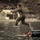U.S. service members train in French Desert Commando Course in Africa A person in camouflage military uniform is captured mid-air leaping from a small wooden dock or platform into or over water. Several other individuals are visible in the water below, appearing to be swimming or treading water near a rubber tire. The setting appears to be a military training exercise near a rocky shoreline with small structures visible in the background.