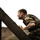 U.S. service members train in French Desert Commando Course in Africa A man wearing camouflage military-style clothing is climbing or scaling a large dark wooden or metal inclined structure. He appears wet, suggesting rain or water exposure, and his expression shows physical exertion. The image has a dramatic, high-contrast cinematic quality with a muted, overcast background.