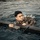 U.S. service members train in French Desert Commando Course in Africa A U.S. Army soldier in full camouflage uniform is in open water, gripping a wooden plank or dock structure while partially submerged. The soldier appears to be exerting significant physical effort, likely during a water training exercise or obstacle course. The expression on the soldier's face suggests strain and determination.