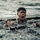 U.S. service members train in French Desert Commando Course in Africa A young man is submerged in open water, emerging with his mouth open and an expression of intense exertion or distress. He appears to be gripping a wooden or metal structure partially submerged beside him. A second individual is visible in the blurred background, also in the water.
