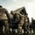 U.S. service members train in French Desert Commando Course in Africa A group of military personnel in multicam/OCP camouflage uniforms are working together to carry or maneuver a blue inflatable rubber assault boat (RHIB or Zodiac-type craft) on a rocky shoreline. The scene appears to be a training exercise involving watercraft operations near a body of water. A large military vehicle or landing craft is visible in the background.