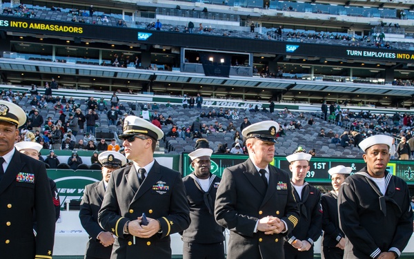 A group of U.S. Navy personnel in dress uniforms are standing on the sideline of an NFL football field, appearing to participate in a military appreciation ceremony. The setting is MetLife Stadium, home of the New York Jets, as indicated by the green Jets branding and scoreboard visible in the background. The stadium is partially filled with spectators, and the scoreboard displays game information.