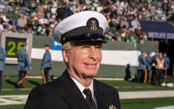 A senior U.S. Navy enlisted officer in dress blues uniform stands on the field at MetLife Stadium during what appears to be a pre-game or halftime military ceremony. He is wearing a white combination cover with a gold anchor insignia and displays multiple rows of service ribbons on his chest. In the background, a color guard or ceremonial unit in blue uniforms is assembled on the field with a packed stadium crowd visible.