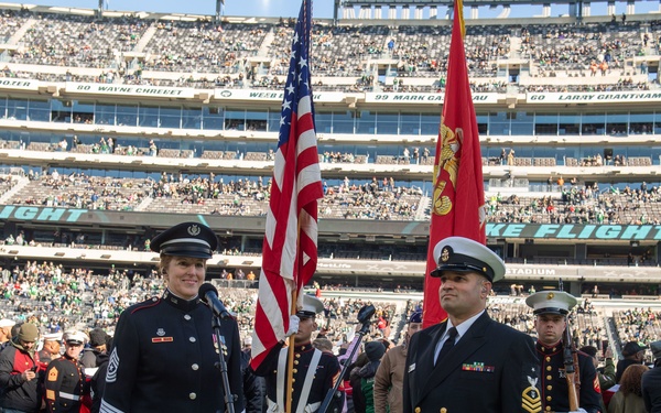 A military color guard ceremony is taking place on the field of a packed NFL stadium, identifiable by the 'Take Flight' signage associated with the New York Jets at MetLife Stadium. Two prominent service members stand at attention in the foreground — a female Army soldier in dress blues and a male Navy senior chief petty officer — flanked by color guard bearers holding the U.S. flag and a Marine Corps flag. The stands are filled with spectators, and a group of veterans or service members is assembled on the field behind them.