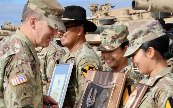 A U.S. Army award ceremony taking place outdoors on a military installation, with soldiers receiving plaques and certificates. Multiple soldiers wearing OCP (Operational Camouflage Pattern) uniforms with 1st Cavalry Division patches are present, holding a 'Secretary of Defense Award' plaque and a framed certificate. Military vehicles, including M1 Abrams tanks with names painted on them, are visible in the background.