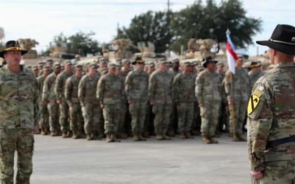 A U.S. Army military formation ceremony is taking place on a paved parade ground. Two senior soldiers wearing distinctive black cavalry Stetson hats are positioned facing a large formation of troops in OCP (Operational Camouflage Pattern) uniforms. Military armored vehicles, likely M1 Abrams tanks, are visible in the background.
