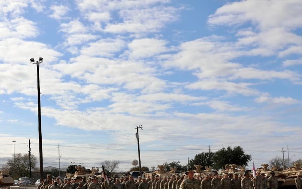 A U.S. Army military formation ceremony is taking place on a large paved parade ground or motor pool area. Soldiers in Army Combat Uniforms (ACU/OCP pattern) are standing in organized ranks while an officer or NCO addresses them from the front. Multiple M1 Abrams main battle tanks are visible in the background, suggesting this is an armored unit.