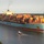A large Maersk Line container ship is navigating a river channel, heavily loaded with multicolored shipping containers stacked several layers high. A small pilot or escort vessel is visible in the water nearby, dwarfed by the massive cargo ship. The scene appears to be at a port city, with urban buildings and tree-lined shores visible in the background.