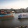 A large Maersk container ship loaded with multicolored shipping containers passes a classic white multi-deck riverboat on a wide river, with a city skyline visible in the background. The scene captures a striking contrast between heavy industrial cargo shipping and leisure/tourism river travel. The setting appears to be the Savannah River near Savannah, Georgia, based on the distinctive skyline featuring church steeples and historic architecture.