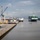 A busy commercial port facility photographed from an elevated vantage point, showing a large container ship approaching or departing along a river channel. The port features multiple cranes, a long pier/wharf structure, and industrial infrastructure in the background. A second smaller vessel is visible mid-channel near the container ship.