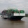 A large Evergreen Line container ship named 'Ever Fortune' is navigating a wide river channel, heavily loaded with stacked shipping containers in green, red, and brown colors. The vessel is being assisted by at least one tugboat visible on its port side. The scene appears to be near a port facility, with green treelines visible on both banks under an overcast sky.