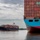 The image shows two large container ships on a wide waterway under an overcast sky. The foreground vessel is the 'Arnold Maersk' registered in Svendborg, featuring a distinctive blue and red hull loaded with hundreds of multicolored shipping containers. A second, smaller container ship is visible in the background, accompanied by a tugboat.