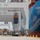 A large container ship named 'COSCO SHIPPING ROSE' is docked at a commercial port terminal. Ship-to-shore cranes are actively loading or unloading shipping containers, with one container visibly suspended mid-air. The terminal yard shows multiple vehicles and equipment operating along the dock.