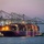 A large ONE (Ocean Network Express) container ship is docked at a port terminal during dusk or dawn, illuminated by artificial lighting. The vessel is heavily loaded with multicolored shipping containers stacked multiple layers high. Multiple large ship-to-shore gantry cranes are positioned over the ship, suggesting active or recent loading/unloading operations.