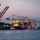 A nighttime/dusk port scene showing two large container ships docked at a busy cargo terminal. The left vessel is clearly marked 'EVERGREEN' and the right vessel appears to be a pink/magenta 'ONE' (Ocean Network Express) ship, both loaded with stacked multicolored shipping containers. Multiple large ship-to-shore gantry cranes are positioned over the vessels, illuminated by port lighting against a purple twilight sky.