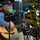 An older man wearing a green face mask, glasses, and a dark baseball cap is seated and playing an acoustic guitar into a microphone. He is performing in front of a decorated Christmas tree adorned with ornaments, tinsel, stockings, and lights. A small amplifier and a music stand are visible nearby, suggesting a live holiday performance.