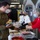 A military service member in camouflage uniform and olive fleece is being served food at what appears to be a holiday meal event, likely on a military base. Volunteers wearing hair nets, masks, and festive accessories are serving food from a long table covered with various dishes in aluminum trays and containers. All individuals are wearing face masks, suggesting this event took place during the COVID-19 pandemic period.