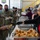 A military meal service event taking place inside what appears to be an aircraft hangar, where civilian volunteers are serving food to uniformed military personnel. All individuals are wearing face masks, suggesting this occurred during the COVID-19 pandemic period. A State Farm volunteer in a red polo shirt and hair net is serving dinner rolls to a soldier identified by name tape as 'Hoskins'.
