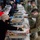 A food service line is being operated in what appears to be an indoor facility, where civilian volunteers in hair nets and masks are serving food to military personnel in camouflage uniforms. Multiple chafing dishes with various foods are arranged along a white-tablecloth-covered serving table. All individuals are wearing face masks, consistent with COVID-19 pandemic-era protocols.