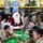 A holiday meal event is taking place inside a military hangar at Dover AFB, Delaware, featuring personnel from the 436th/512th Aircraft Generation Squadron and Aircraft Maintenance Squadron. Military personnel in OCP (Operational Camouflage Pattern) uniforms are seated at green-covered tables eating a holiday meal, while a person dressed as Santa Claus mingles with the group. The event appears to be a Christmas or holiday morale event, with some attendees wearing face masks consistent with COVID-era protocols.