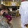 A military service member in U.S. Army/Air Force camouflage uniform holds a young toddler while interacting with a person dressed as Santa Claus inside what appears to be a military hangar or maintenance facility. Both the service member and child are wearing face masks, suggesting this event took place during the COVID-19 pandemic. A decorated Christmas tree with wrapped presents is visible in the background.