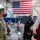 A military holiday event taking place inside what appears to be a hangar or large military facility, decorated for Christmas. A uniformed U.S. Air Force service member and a civilian are conversing with a person dressed as Santa Claus, all wearing face masks consistent with COVID-19 precautions. A large American flag is prominently displayed on the wall in the background.