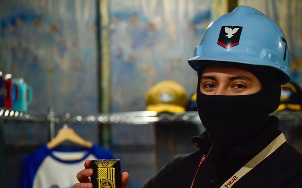 A person wearing a light blue hard hat with a military insignia and a black balaclava is holding up a Zippo lighter in what appears to be a ship's store or retail area. The individual is wearing a lanyard that reads 'Ship's Store' and appears to be a U.S. Navy sailor displaying merchandise. The background shows clothing racks with various items including caps and shirts.