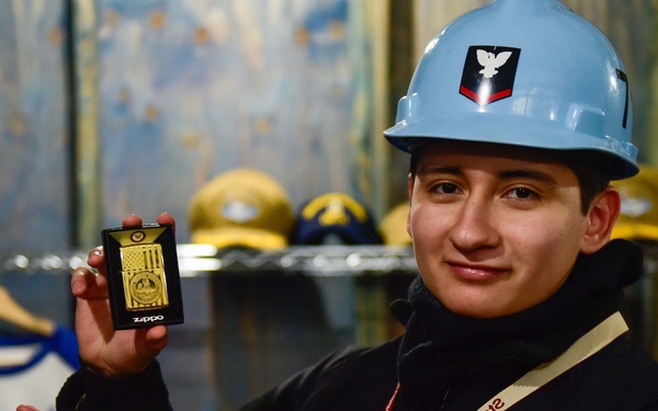 A young person wearing a light blue hard hat with a military insignia is holding up a gold Zippo lighter toward the camera with a smile. They are wearing a black jacket and a red lanyard that reads 'Theodore Roosevelt' and 'Ship's Store'. The background shows a retail merchandise area with baseball caps, shirts, and other items on display.