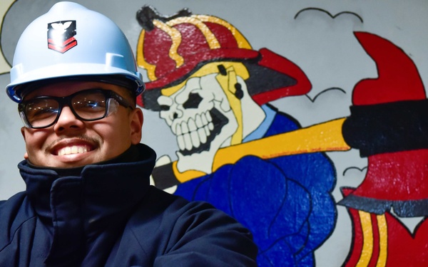 A smiling man wearing a light blue hard hat and dark jacket stands with arms crossed in front of a painted mural. The mural depicts a skeleton dressed as a firefighter holding an axe, wearing a red helmet and boots. Text visible in the background includes '45-252 DIV' suggesting a military or industrial setting.