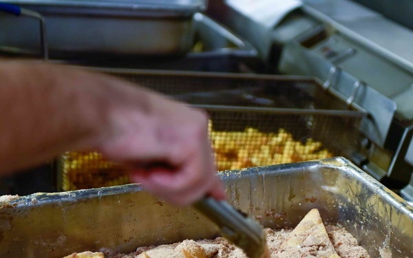 A person is working in a commercial kitchen, coating fried tortilla chips in a seasoning or spice mixture inside a stainless steel hotel pan. The process appears to involve tossing or mixing the chips with a powdered coating using tongs. A second pan with what appears to be waffle-cut fries or chips is visible in the background.