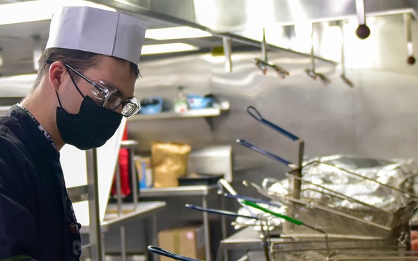 A cook or food service worker wearing a white chef's hat, black face mask, and glasses is working at a commercial kitchen fry station. They are using green-handled tongs to handle breaded food items in a stainless steel pan. The setting appears to be a large institutional or military galley kitchen based on the scale and equipment visible.