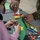 Celebrating the New Year at Task Force Liberty A child wearing a face mask and pink hoodie is doing a craft project with an adult in military camouflage uniform. They are working together on a green paper craft decorated with foam stickers including a turtle and star, using Elmer's School Glue. The table is covered with various art supplies including markers, pipe cleaners, pom-poms, and glitter decorations.