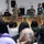 Celebrating the New Year at Task Force Liberty A military officer in camouflage uniform speaks into a microphone on a stage before a seated audience inside a large tent or temporary facility. Several civilians wearing safety vests and face masks stand nearby, suggesting an organized briefing or information session. An Afghan flag is visible on the decorated table, indicating this may be a gathering related to Afghan refugee operations.