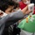 Celebrating the New Year at Task Force Liberty A young child wearing a white face mask is engaged in an arts and crafts activity, applying glue from a bottle onto a green sheet of paper. The craft project includes colorful pom-poms and a small white paper turtle cutout being arranged on the green background. Other masked children are visible in the blurred background, suggesting a group craft session.